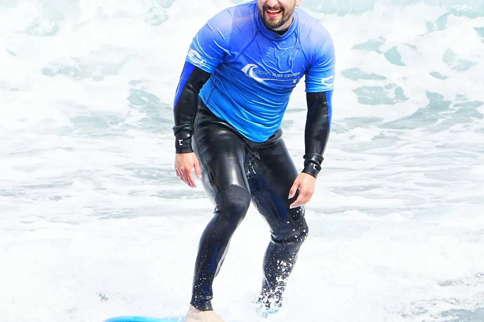 Un surfista sorridente in lycra Azores Surf Center prende un’onda su una tavola blu