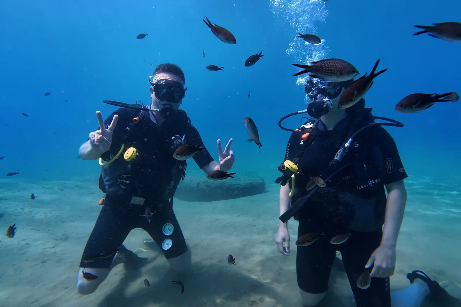 Due subacquei di Divers Club Crete inginocchiati sul fondo sabbioso, circondati dai pesci, fanno il segno della pace