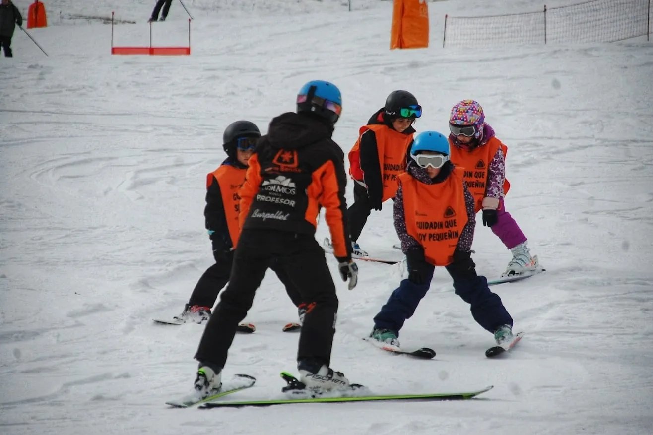 An instructor from the Valle de Benás ski school leading a group of young skiers in orange high-visibility bibs down a beginner slope, watching their stance as they come