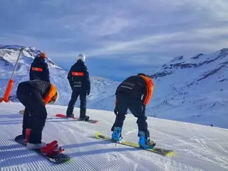 Three instructors in the school's black and orange jackets standing at the top of a freshly groomed run at first light, the snow-covered Pyrenees spread out below them