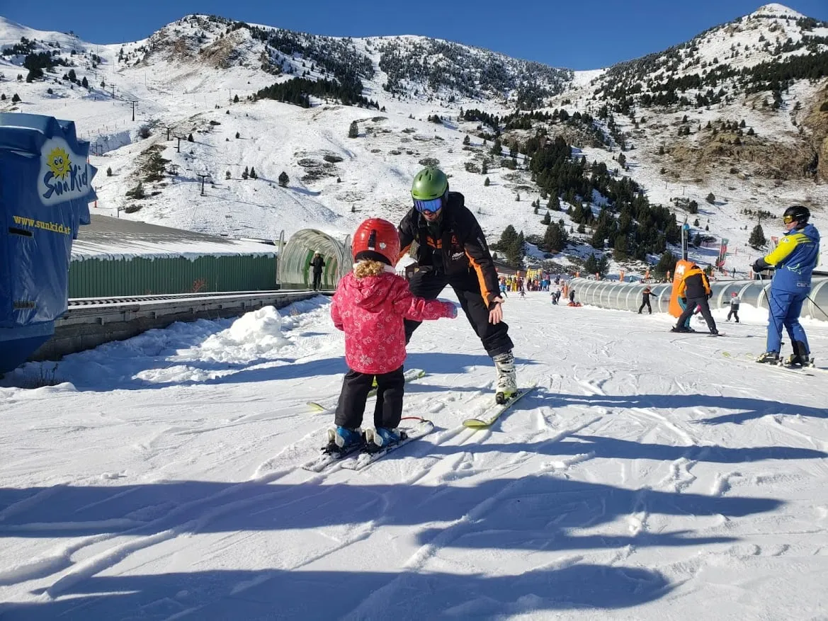 An instructor guiding a small child in a pink jacket through their first turns beside the beginners' carpet lift, with the wooded slopes of Cerler rising behind