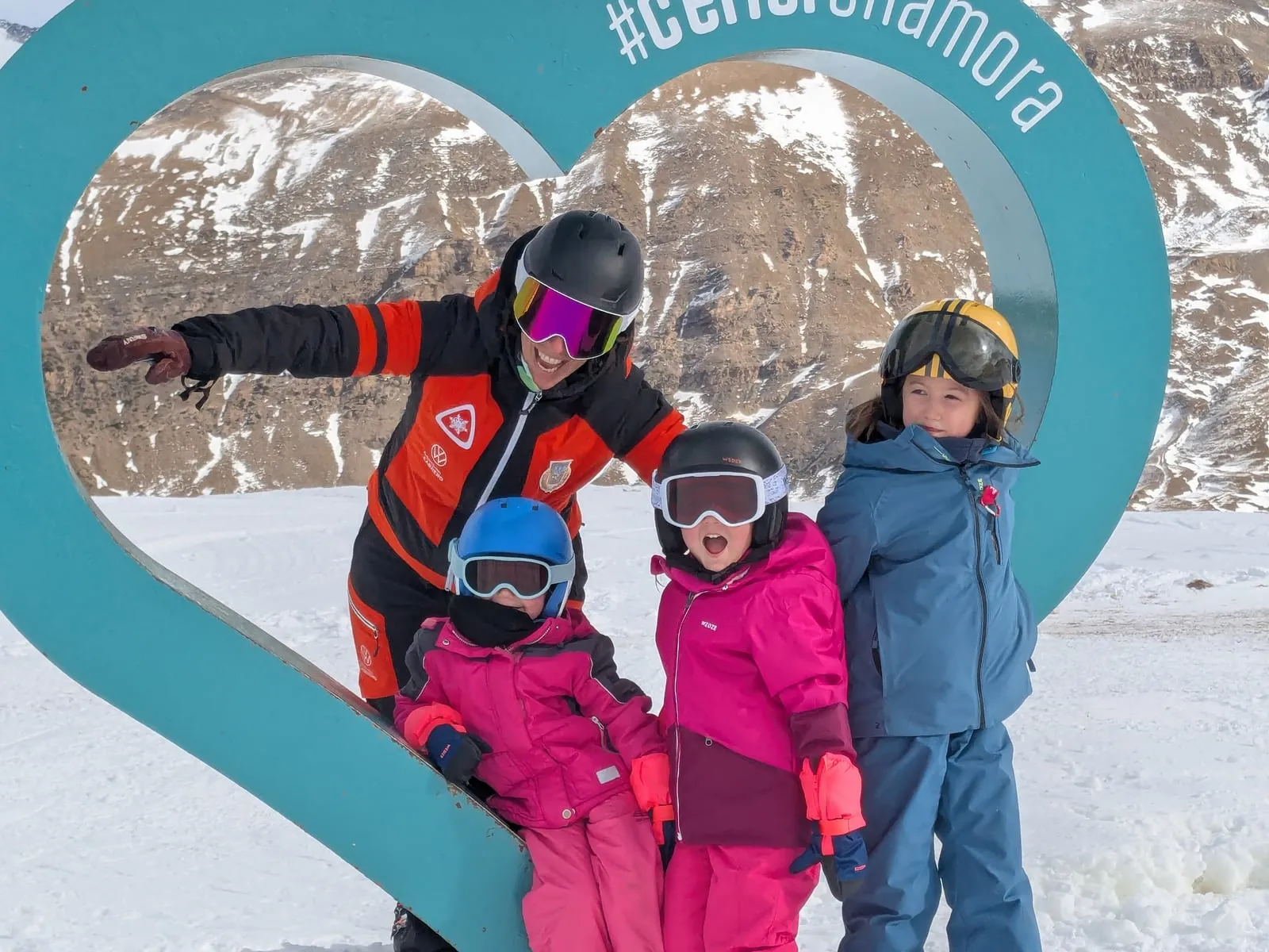 An instructor with three young pupils in front of the turquoise heart-shaped sign at Cerler, snow and rocky ridges behind them