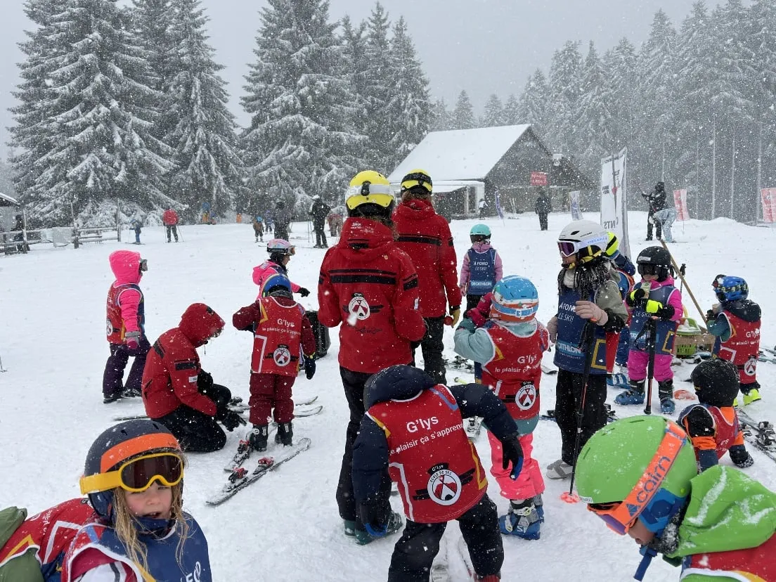 Des moniteurs de l’école de ski G'lys en vestes rouges rassemblant des enfants en dossards rouges, skis aux pieds, sous la neige qui tombe, le chalet de l’école et ses drapeaux parmi les sapins derrière