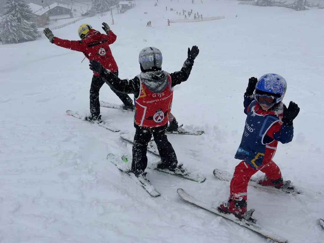 Un moniteur et deux jeunes élèves à ski prenant la pose, bras levés, sur une pente enneigée, avec un petit remonte-pente et des chalets dans la neige en contrebas