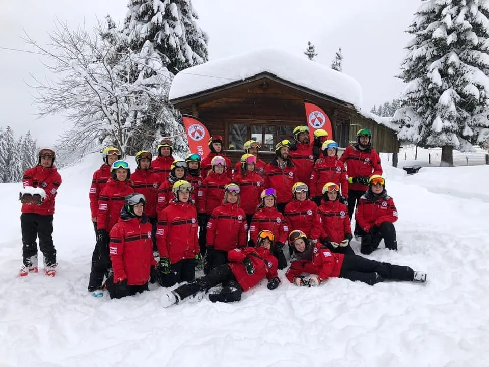 L’équipe de moniteurs de l’école de ski G'lys, en vestes rouges et casques assortis, posant dans la neige épaisse devant un chalet en bois enneigé orné des drapeaux de l’école