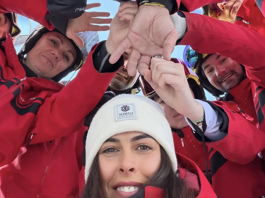 Des moniteurs de l’école de ski G'lys en vestes rouges, les mains empilées en cercle, photographiés d’en bas sur fond de ciel bleu