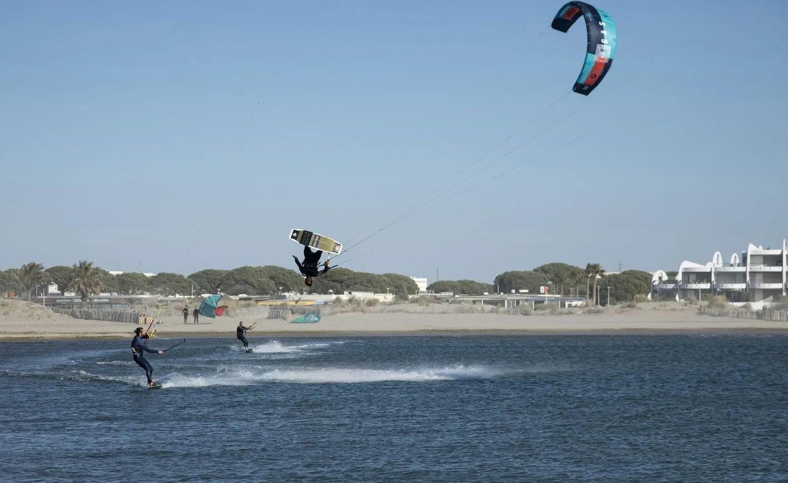 A Seb Garat Ride Academy kitesurfer launching a big jump over the water with two riders below and the coast behind