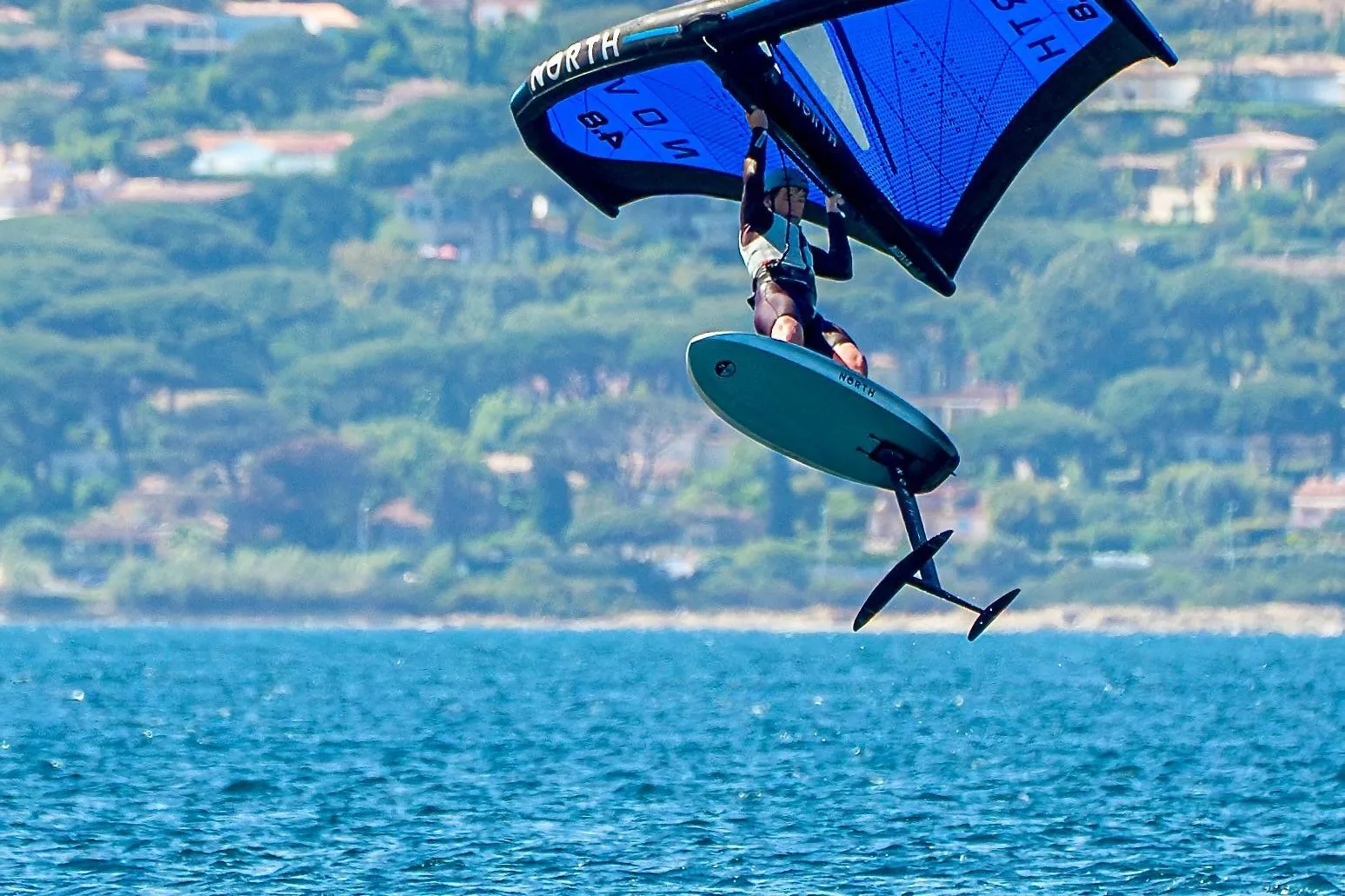 Wing foiler jumping clear of the water with the board lifted high over the bay of Saint-Tropez
