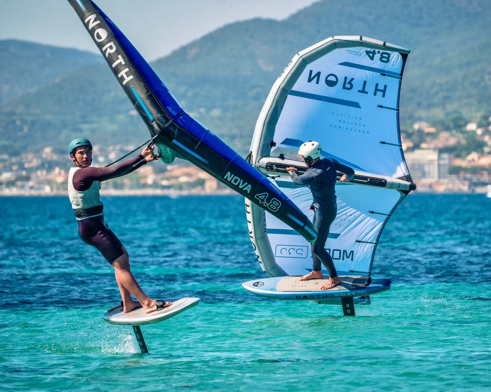 Two wing foilers riding side by side on the bay of Saint-Tropez with the hills of the Riviera behind