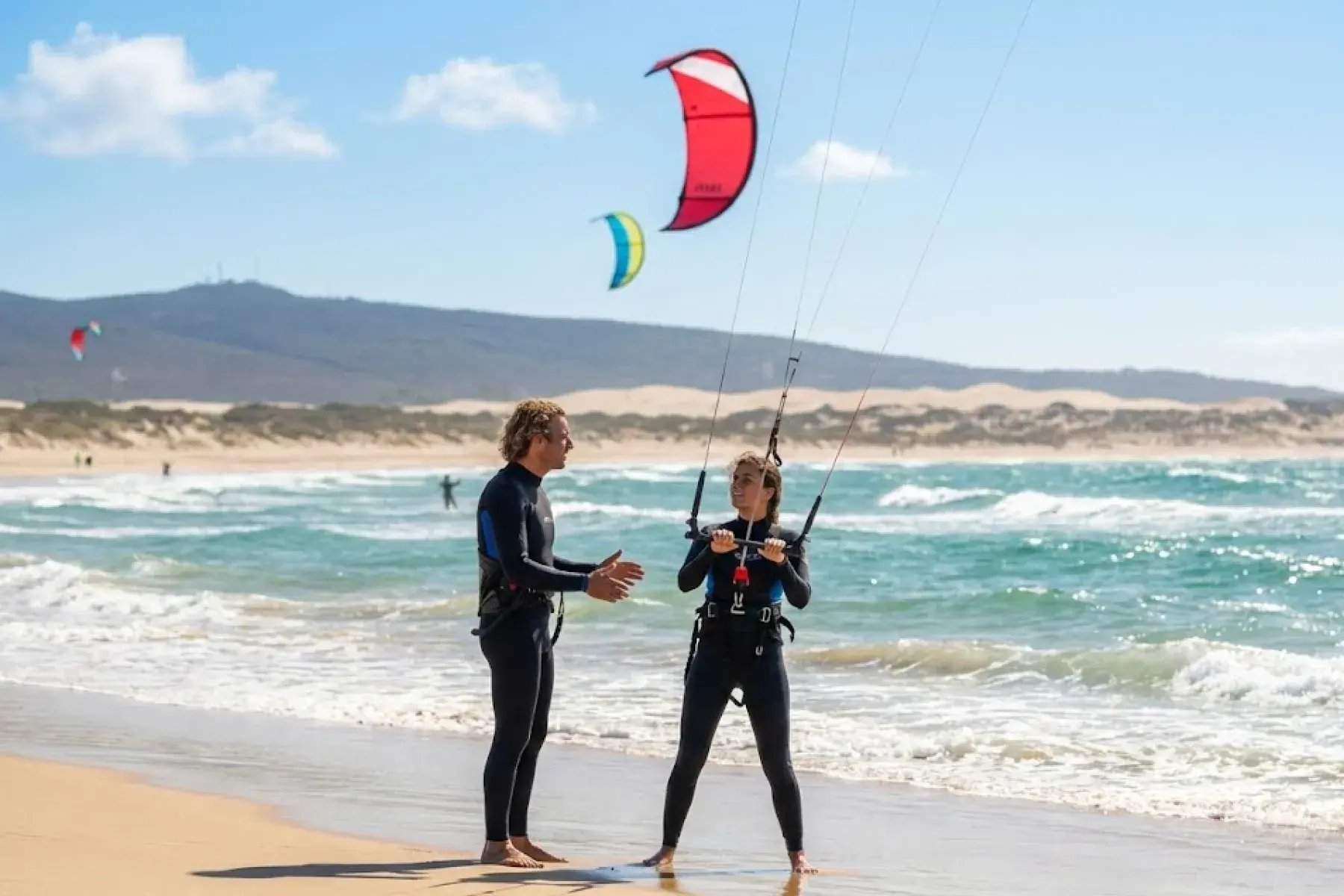 Instructor and student on a sandy beach during a kitesurfing lesson with kites flying overhead