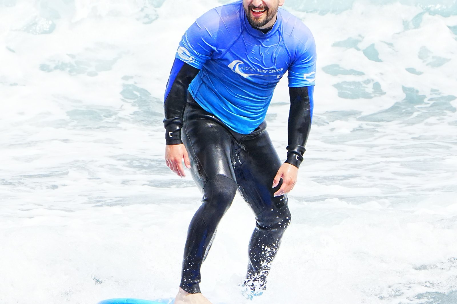 Un surfista sorridente in lycra Azores Surf Center prende un’onda su una tavola blu