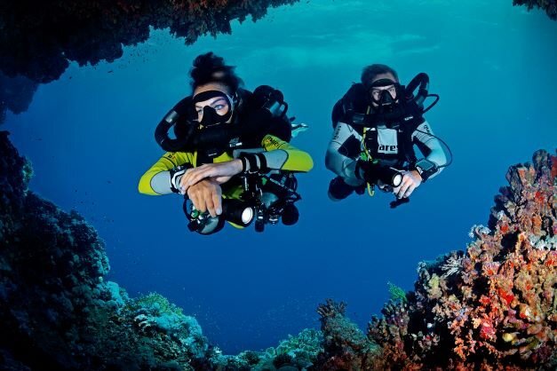 Due subacquei di Buceo Cabo La Nao avanzano sotto una sporgenza rocciosa ricoperta di gorgonie colorate