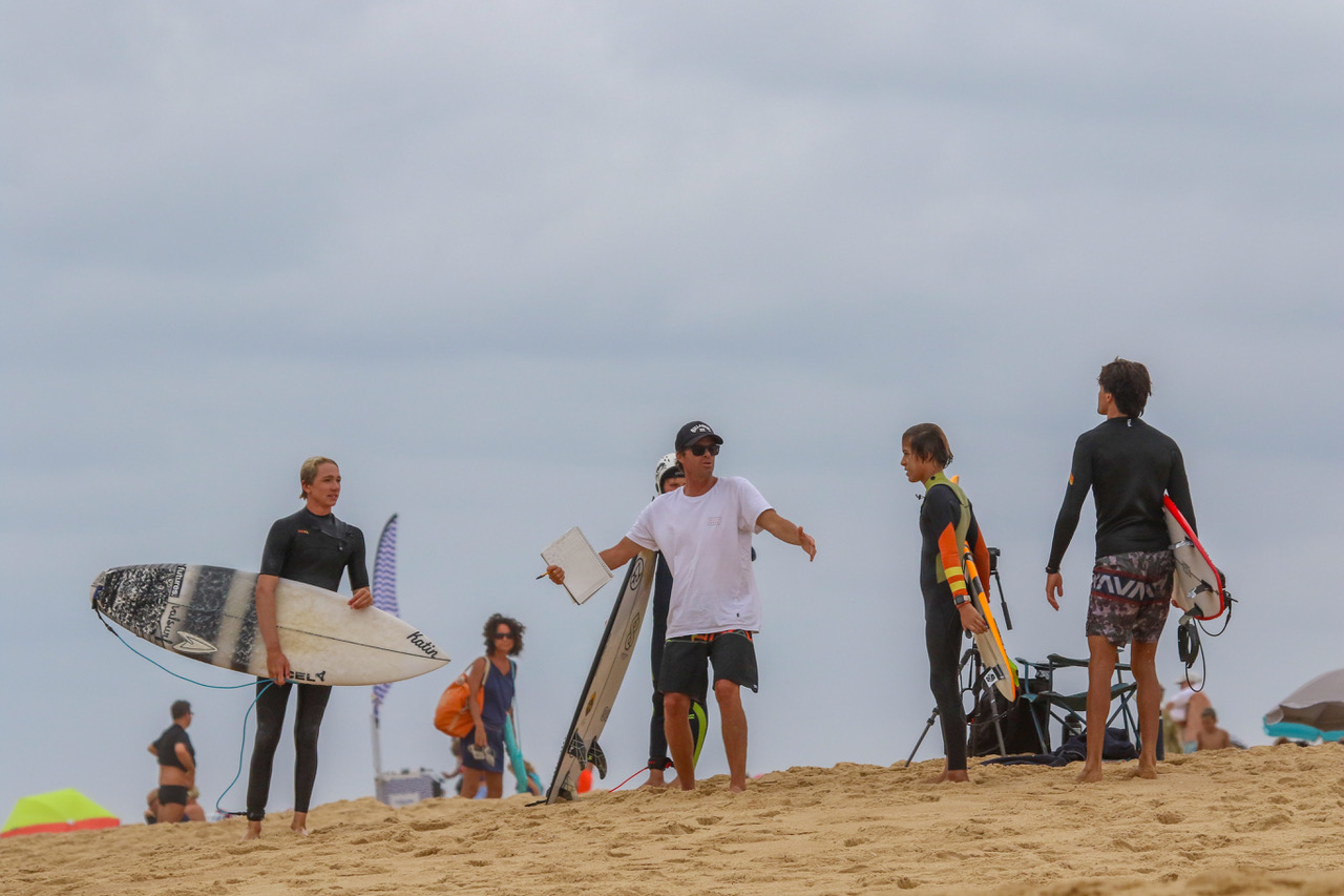 Didier Piter fa un briefing agli studenti di surf in spiaggia con un clipboard mentre tengono le tavole