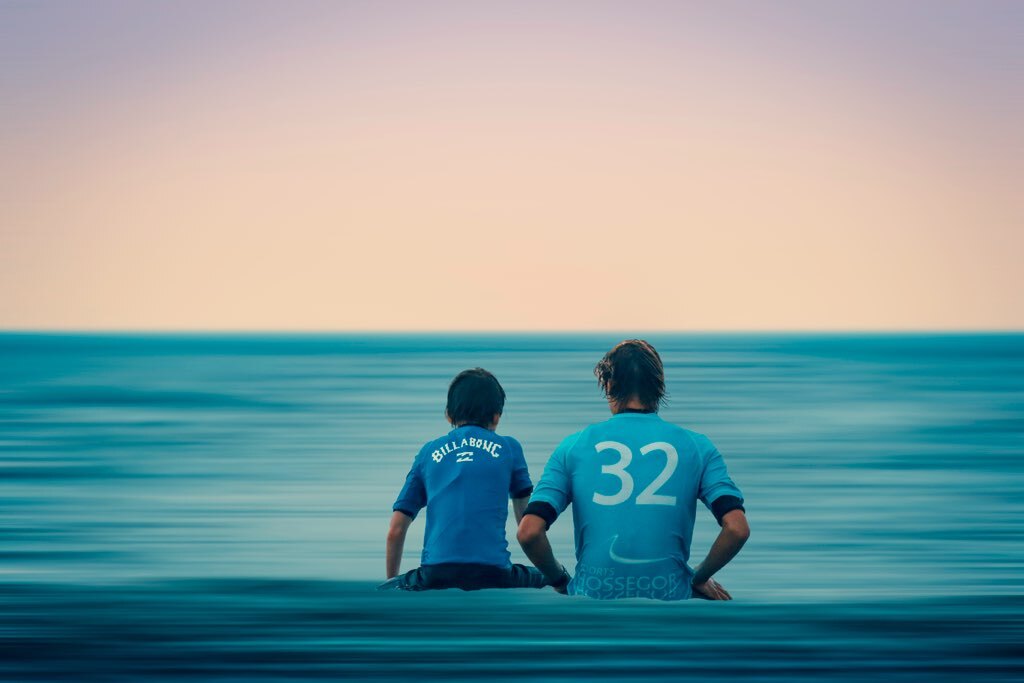 Surf instructor and young student sitting together in the ocean looking out at the horizon at sunset in Hossegor