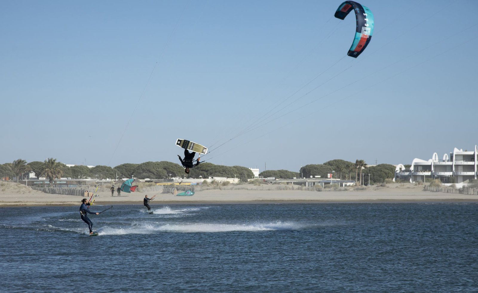 Un kitesurfista di Seb Garat Ride Academy salta in alto sopra l’acqua, con due rider sotto e la costa alle spalle