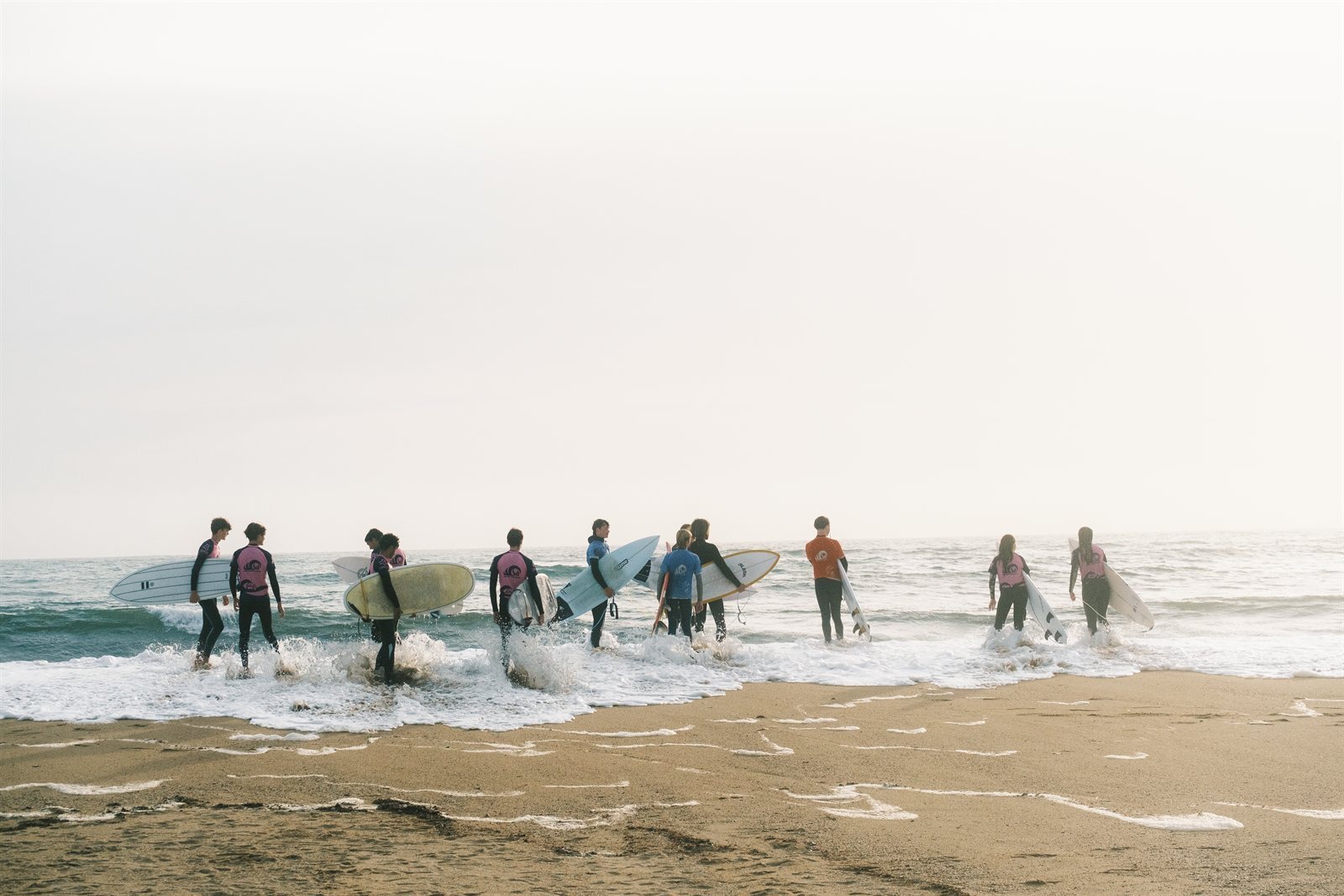 Group of WSA Guidel surf students in wetsuits carrying surfboards into the ocean at Guidel Plages