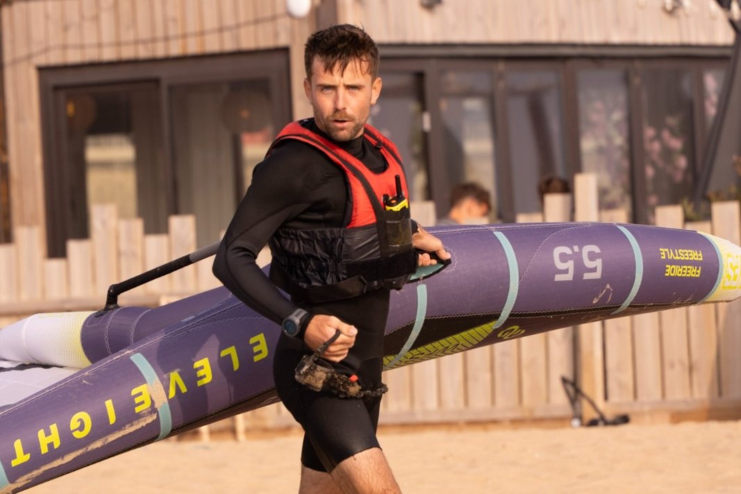 Un istruttore Yagga in muta porta una tavola da wingfoil sulla spiaggia, davanti alla base